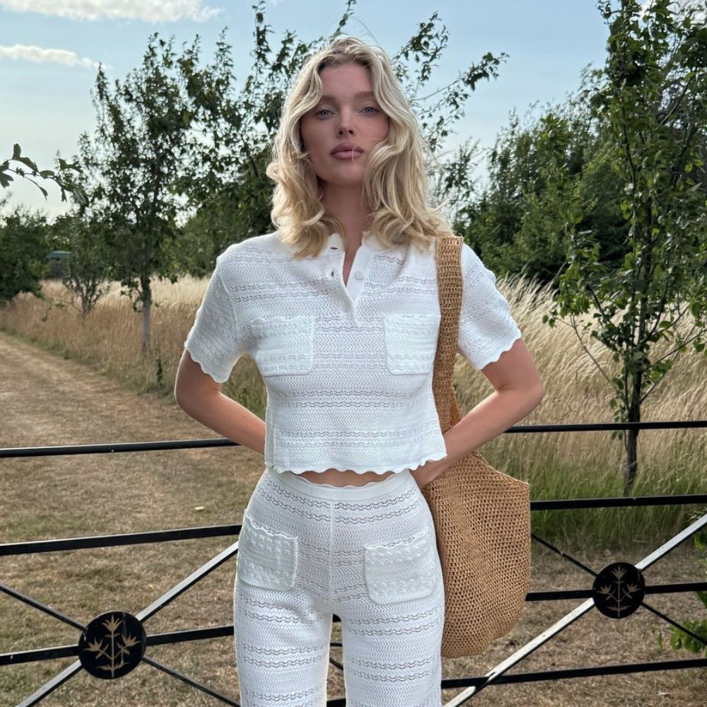 Elsa Hosk stands outside wearing a white crochet top and trouser co-ord with a brown woven bag and brown sandals.