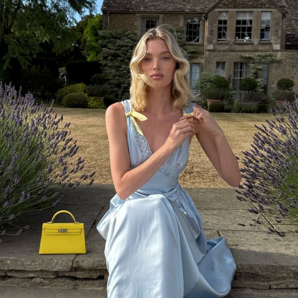 Elsa Hosk sits outside in a garden in front of an English countryside house wearing a blue satin maxi dress with yellow bows on the arms and a yellow Hermes bag.